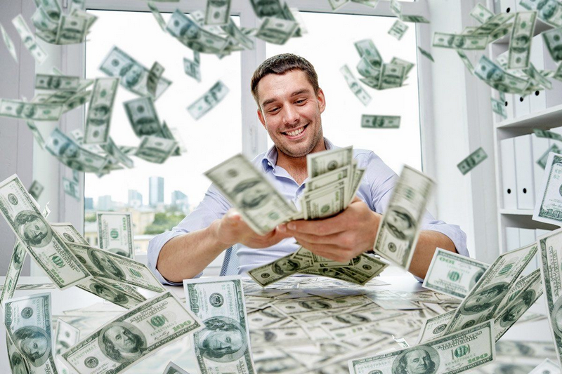 A man smiling at a table, surrounded by flying dollar bills.