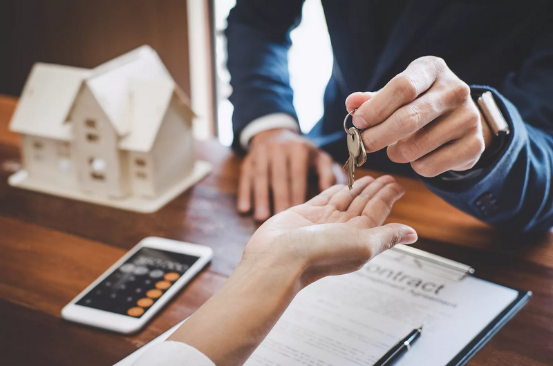 A person hands over house keys to another person across a wooden table with a contract, phone, and a house model.