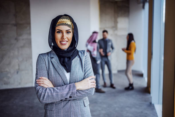 Woman in business attire smiling confidently with colleagues discussing in the background, representing success in real estate.