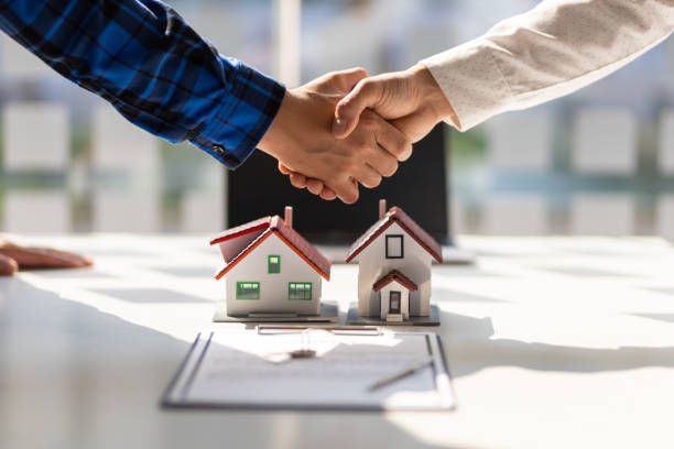 A handshake over miniature house models and a contract signifies closing an apartment purchase deal.