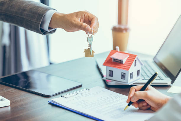 Person handing over keys and another signing documents with a small house model on the desk.